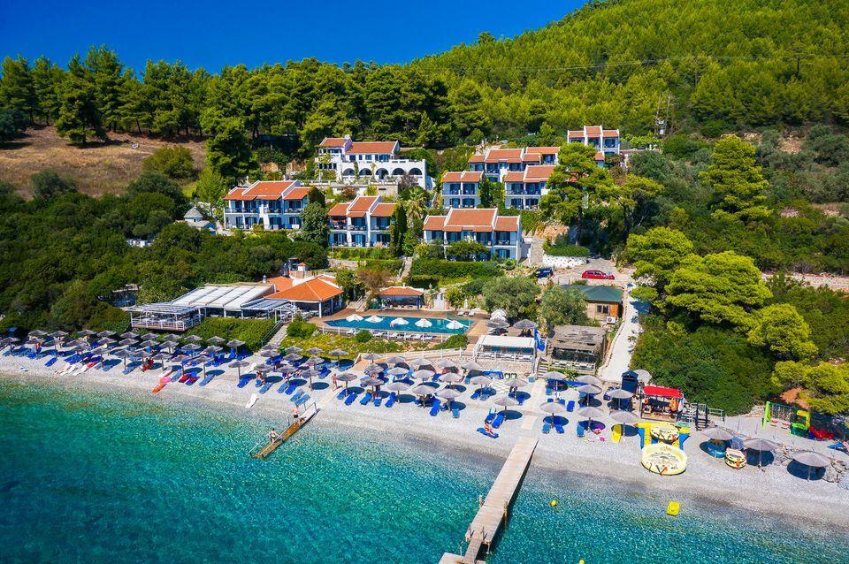 Adrina Beach beside Adrina Beach Hotel on Skopelos, with clear Aegean water and forested hills beyond.