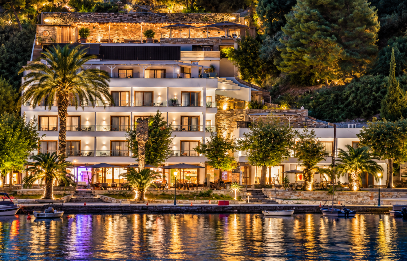 Natura Luxury Boutique Hotel in Loutraki, Skopelos, illuminated at night above the waterfront, with white buildings, stone terraces, palm trees, moored boats and warm lights reflected in the calm water.