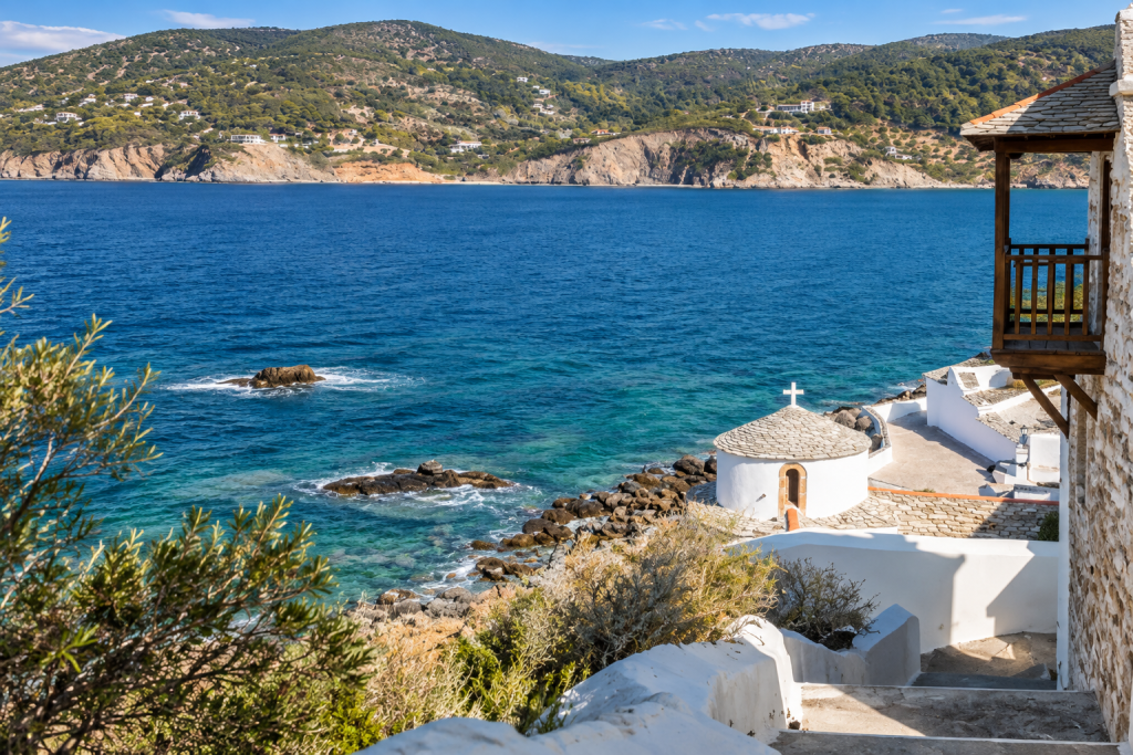 Whitewashed coastal chapel and stone steps overlooking the blue Aegean Sea, with rocky shoreline, wooded hills and scattered villas across the bay.