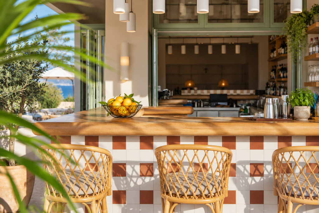 Boutique hotel bar with a wood-topped tiled counter, woven rattan stools, pendant lights and open windows overlooking greenery and the Aegean Sea.