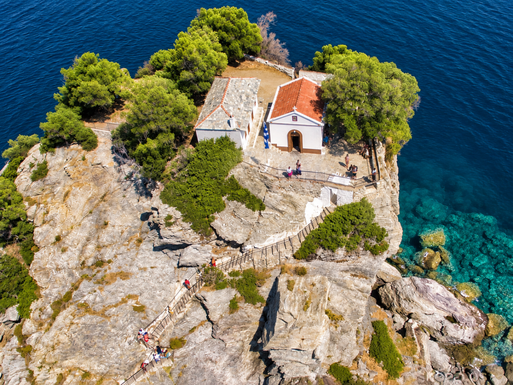 Aerial view of a small rocky islet off Skopelos with a white chapel, terracotta roof, pine trees, stone paths and railings surrounded by deep blue Aegean water.