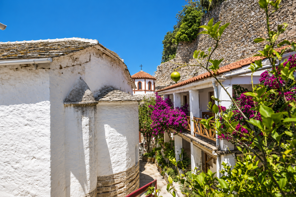Sunlit narrow lane in Mount Palouki, Skopelos, lined with whitewashed buildings, a small stone chapel, terracotta roofs, a hillside stone wall and vibrant bougainvillea.