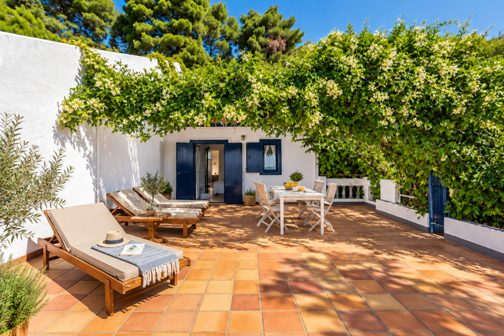 Sunlit outdoor terrace at Adrina Beach Hotel on Skopelos, with terracotta tiles, white walls, blue shutters, shaded loungers, dining furniture and flowering greenery.