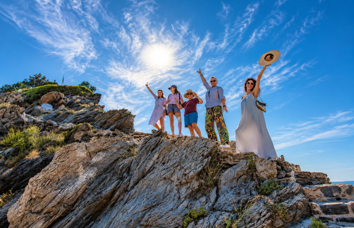 Group of visitors standing on a rugged coastal rock formation on Skopelos, raising their arms beneath a bright blue sky with wispy clouds and Mediterranean vegetation.