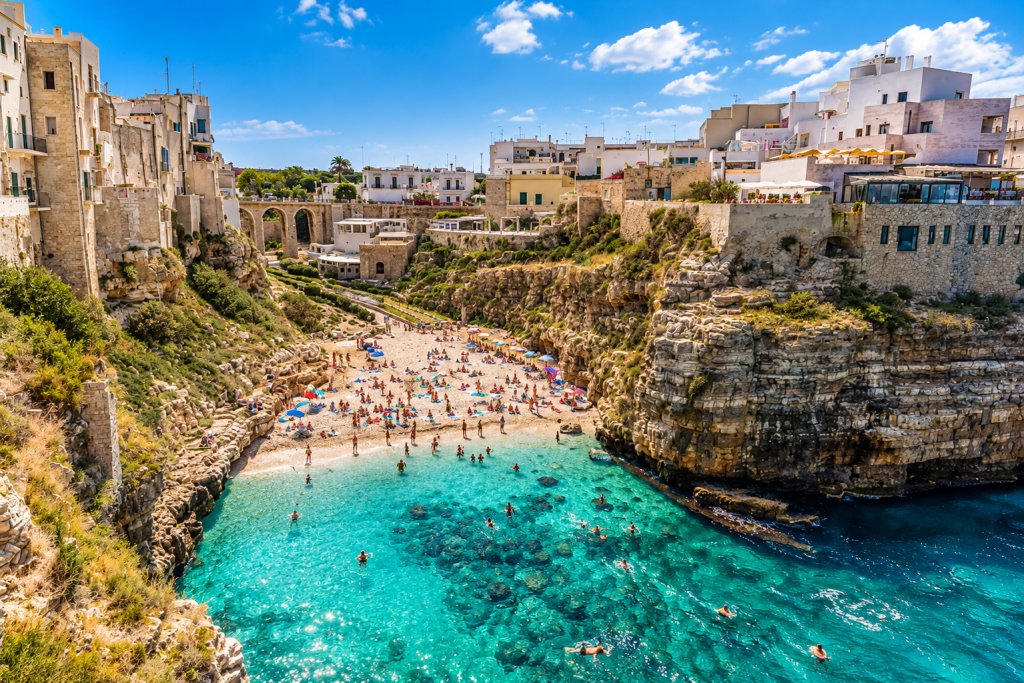 Turquoise coastal cove in southern Italy, surrounded by rugged limestone cliffs and whitewashed buildings, with a busy pebble beach and swimmers in the clear water.