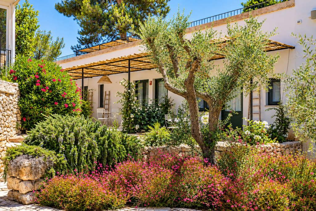 Masseria Fontanelle courtyard with white stone buildings, shaded pergolas, an olive tree and Mediterranean flowering gardens.