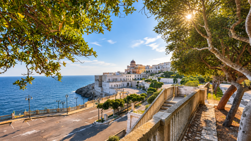 Coastal terrace overlooking a whitewashed Mediterranean town, deep blue sea and rocky shoreline, framed by leafy trees and stone steps.