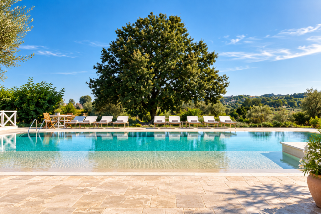 Swimming pool at Leonardo Trulli Resort near Locorotondo, surrounded by sun loungers, Mediterranean gardens, olive trees and rolling countryside under a clear blue sky.