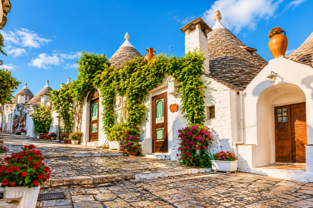 Whitewashed trulli with conical stone roofs lining a cobbled lane in Alberobello, framed by climbing vines, colourful flowers and terracotta pots in warm afternoon light.