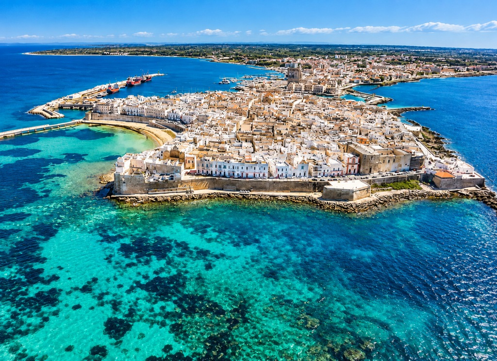Aerial view of Otranto’s historic peninsula surrounded by clear turquoise and deep-blue Adriatic water, with dense pale-stone buildings, harbour areas, beaches and waterfront fortifications.