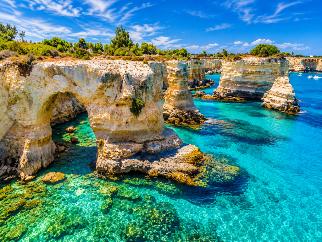 Layered limestone cliffs and sea arches along the Puglian coast, rising above clear turquoise Adriatic water with submerged rocks and Mediterranean vegetation.