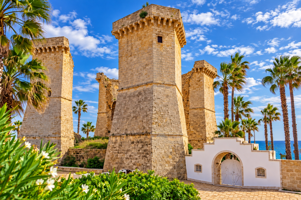 Historic limestone coastal fortress with tall square towers, palm trees and flowering Mediterranean gardens, with the blue sea visible beyond under a partly cloudy sky.