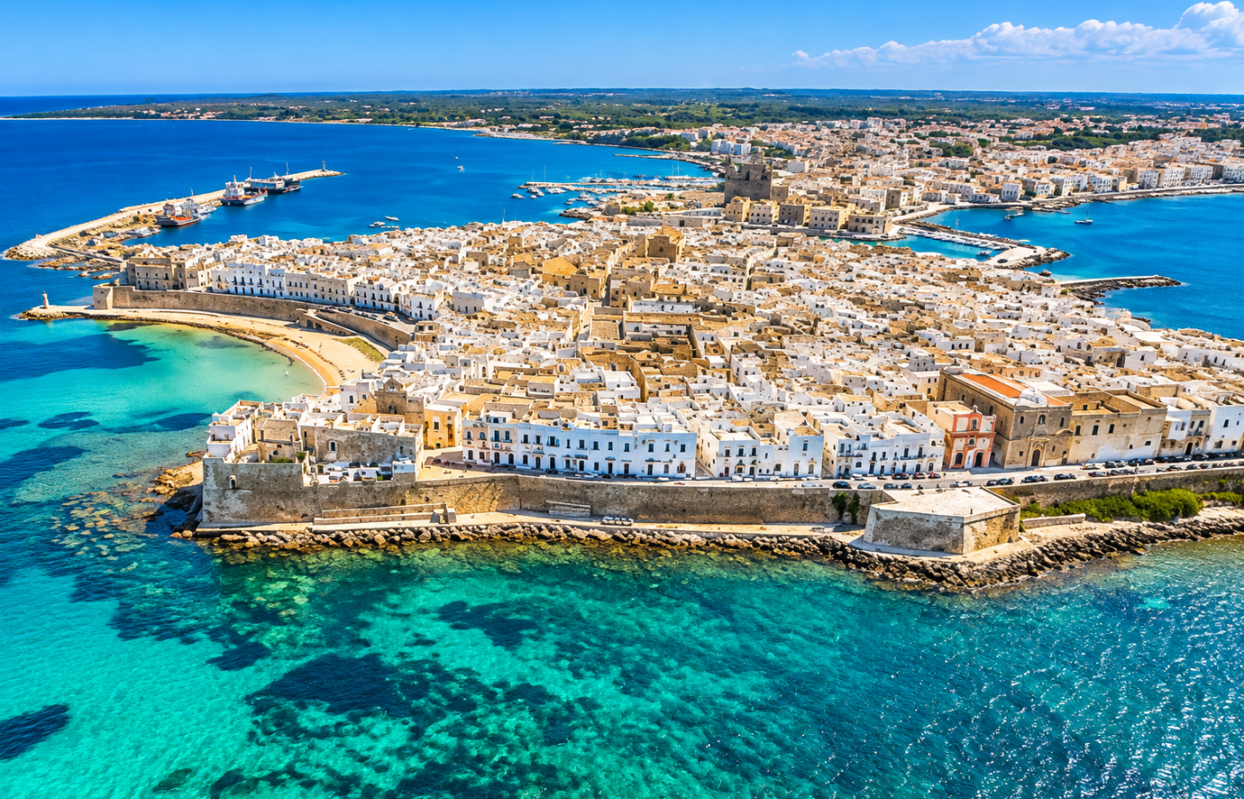 Aerial view of Gallipoli’s historic fortified peninsula, with dense pale-stone buildings, harbour walls, beaches and vivid turquoise Ionian Sea surrounding the old town.
